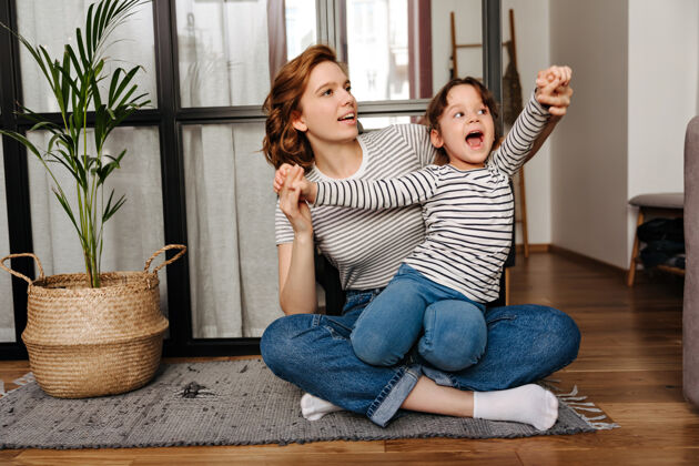 拥抱穿着条纹t恤的红发女人抱着女儿 和她坐在客厅的地板上玩耍童年可爱女性