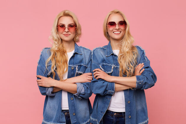 女士积极的年轻迷人的长发金发女人 戴着红色太阳镜 微笑着看着镜头 双手交叉着站在粉色背景上双胞胎女士姐妹