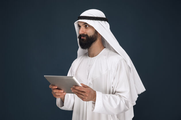 沙特深蓝色墙上的半身阿拉伯沙特商人肖像年轻的男性模特使用平板电脑或小玩意儿商业概念 金融 面部表情 人类情感 技术情绪表情惊喜