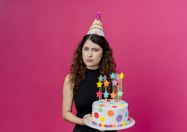 帽子年轻漂亮的女人卷发戴节日帽拿着生日蛋糕不高兴皱眉脸生日派对概念站在粉红色的墙上年轻脸抱着