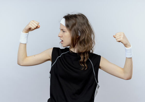 年轻穿着黑色运动服的年轻健身女孩 头上戴着头巾 举起拳头 愤怒的脸站在白色的墙上举起健康头带