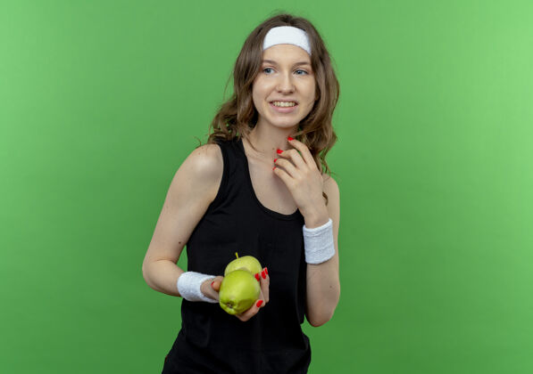 站着身穿黑色运动服的年轻健身女孩 头箍上拿着两个绿色的苹果 站在绿色的墙上欢快地笑着年轻健身抱着