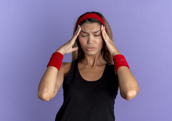 手身穿黑色运动服 头戴红色头巾的年轻健身女孩 站在蓝色的墙上 手碰到头 头痛得厉害看头带站