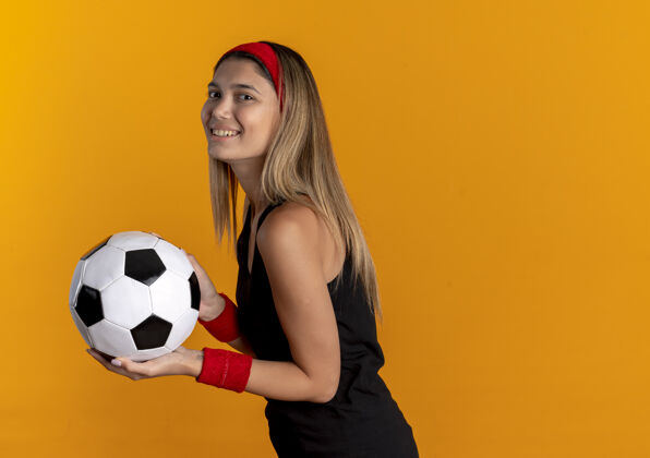 抱身穿黑色运动服 头戴红色头巾 面带微笑地拿着足球站在橘色墙上的年轻健身女孩年轻足球看