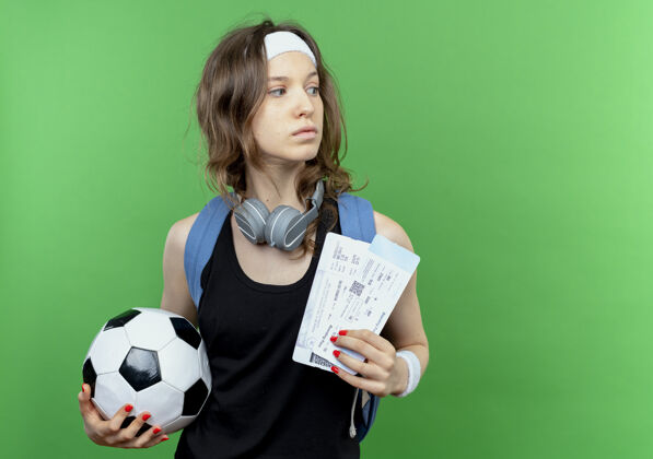 立场年轻的健身女孩穿着黑色运动服 背着背包 戴着头巾 手里拿着足球和机票 站在绿色的墙上困惑地看着一边举行球拼图