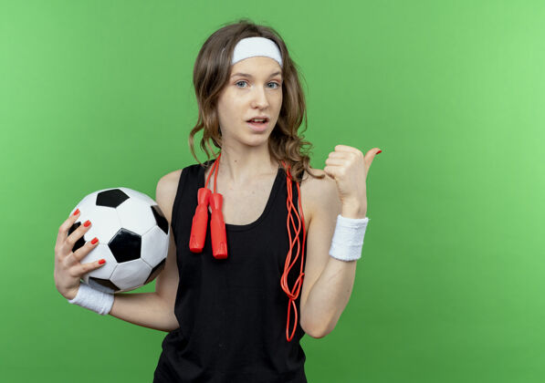 绳子身穿黑色运动服的年轻健身女孩戴着头带 脖子上系着跳绳 手里拿着足球 困惑地指着背站在绿色的墙上周围球足球
