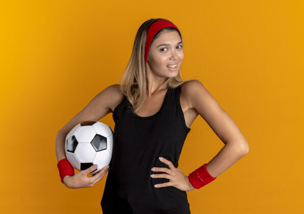 微笑身穿黑色运动服 头戴红色头巾 手持足球 面带微笑的年轻健身女孩站在橙色的墙上看站年轻