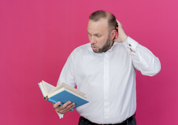 抱着一个留着胡子的男人穿着白衬衫 手里拿着一本打开的书 站在粉红色的墙上困惑地看着它开放男人戴着