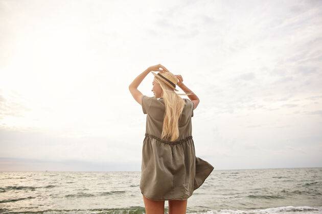 海岸一张明亮的户外照片 年轻的白头女性高举双手戴着草帽 一边欣赏着海边的景色 一边穿着浪漫的绿色连衣裙 背景是海滩沙滩帽子天空
