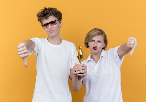 女人年轻的运动情侣站在一起拿着奖杯 竖起大拇指站在橙色的墙上拇指向下表演