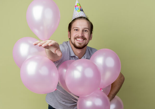 男人戴着节日帽的年轻人举着气球欢庆生日聚会 站在轻薄的墙上兴奋地笑着举行微笑欢呼