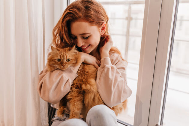 红猫轻松微笑的女孩玩她的毛茸茸的猫室内拍摄的惊人的女士抱着宠物动物积极女模特