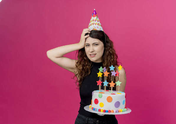 帽子一个年轻漂亮的女人 卷发 戴着节日帽 手里拿着生日蛋糕 头上戴着一只手 站在粉色的墙上 看起来很困惑生日派对的概念头抱着困惑