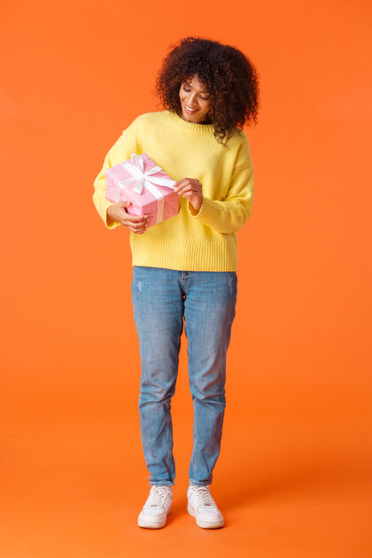 快乐全长垂直拍摄可爱和快乐的生日女孩拆开礼物 微笑的非洲裔美国妇女卷发毛衣 持有粉红色的礼物好奇里面是什么 橙色的墙现在年轻乐趣