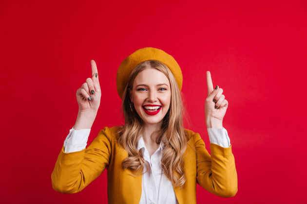 肖像幸福的年轻女子戴着贝雷帽 手指向上在红墙上看到一个迷人的微笑女孩女人情感法国