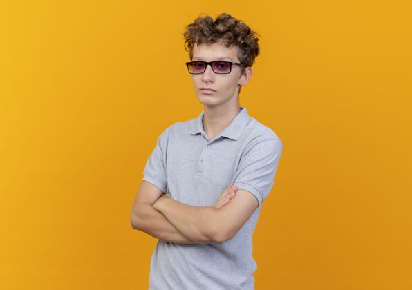 交叉戴着黑眼镜的年轻人 穿着灰色马球衫 严肃的脸 双手交叉放在胸前 站在橙色的墙上脸马球年轻