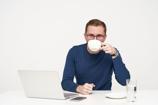 理发摄影棚拍摄的年轻胡须男戴着眼镜 一边喝咖啡 一边用笔记本电脑工作 一边看着相机 穿着蓝色毛衣 坐在白色背景上套头衫男人桌子