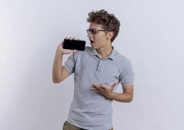 歌手穿着灰色马球衫的年轻人拿着智能手机当麦克风站在白墙上唱歌站立使用手机