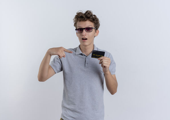 年轻人一个穿着灰色马球衫的年轻人站在白墙上 用手指着信用卡 很惊讶指点表演手指