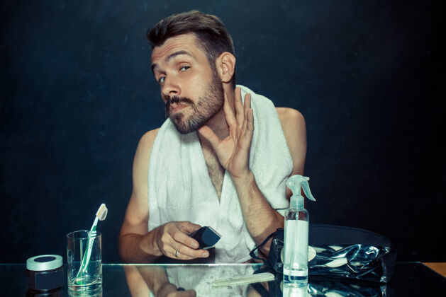 剃卧室里的年轻人坐在镜子前 在家里搔胡子人类的情感和生活方式的概念干净人毛巾