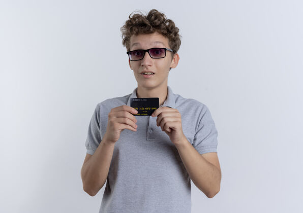显示一个戴着黑眼镜 穿着灰色马球衫 出示信用卡的年轻人站在白色的墙上惊讶不已男人站年轻