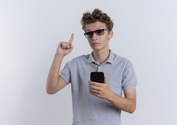 年轻戴着黑眼镜的年轻人 穿着灰色马球衫 手持智能手机 食指露出惊讶的表情 站在白色的墙上 有了新的想法穿马球表演