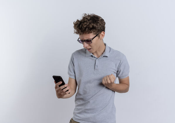 屏幕穿着灰色马球衫的年轻人看着他的智能手机屏幕握紧拳头站在白色的墙上 高兴而兴奋男人握紧站