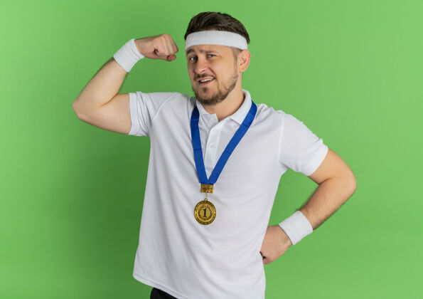 展示身穿白衬衫 头箍和金牌环绕脖子的年轻健身男子举起拳头展示肱二头肌 绿色背景下站着赢家概念拳头衬衫奖牌