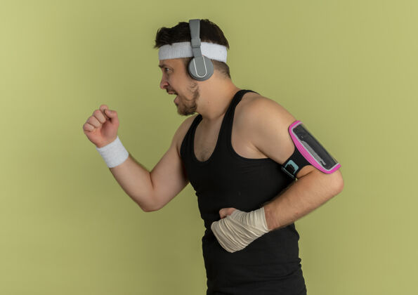 立场戴着头带和耳机的年轻健身男子戴着智能手机袖标站在橄榄色背景下训练臂带男子耳机