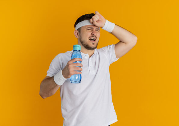 看身穿白衬衫 头箍 拿着一瓶水的年轻健身男子站在橙色背景下 看上去既疲惫又疲惫白色锻炼男人