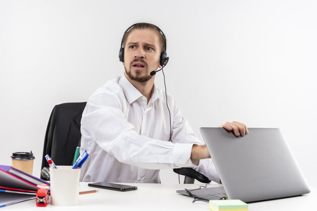 工作穿着白衬衫 戴着耳机 拿着麦克风的帅哥坐在白色背景下的办公桌旁 严肃地看着一旁的笔记本电脑耳机严肃帅气