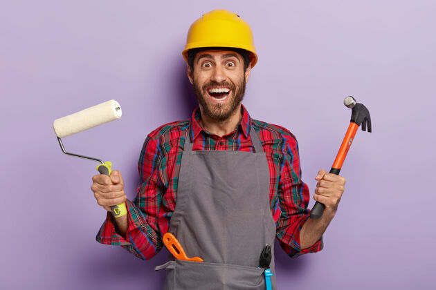 油漆工积极的维修人员拿着锤子和油漆辊 戴着围裙和安全帽 有许多建筑工具 随时准备装修房子快乐的专业服务人员可以修理你公寓里的一切微笑头盔高兴