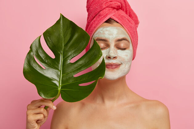 女性健康的欧洲女性用面膜复壮去除毛孔 握住绿叶 闭上眼睛 赤裸的身体 裹在头上的毛巾 造型在室内美容 美容植物姿势化妆品