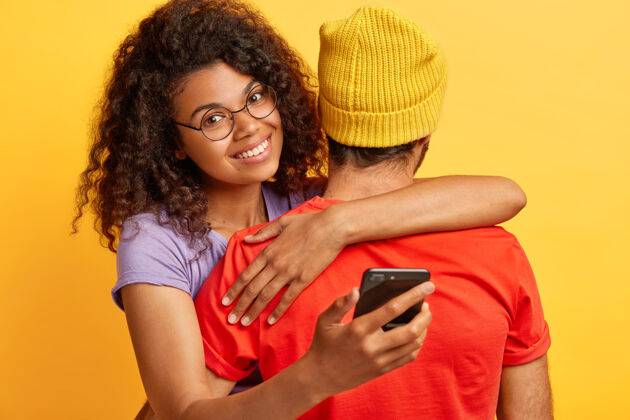 理发快乐的黑皮肤非洲发型女人 戴着圆眼镜 拥抱着戴着黄帽子和红t恤的男人 拿着手机 等待重要的电话人 技术 关系概念设备拥抱满意