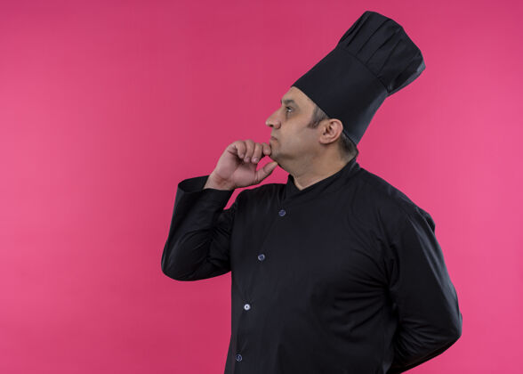 男性男厨师身穿黑色制服 头戴厨师帽 手放在下巴上 面带沉思的表情 站在粉色背景上穿站旁边