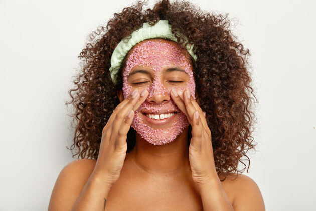 头带快乐美丽的非洲裔美国女人的形象用粉色海盐磨砂膏去皮 抚摸脸颊 光着肩膀靠着白墙个人护理和美容理念年轻化盐健康
