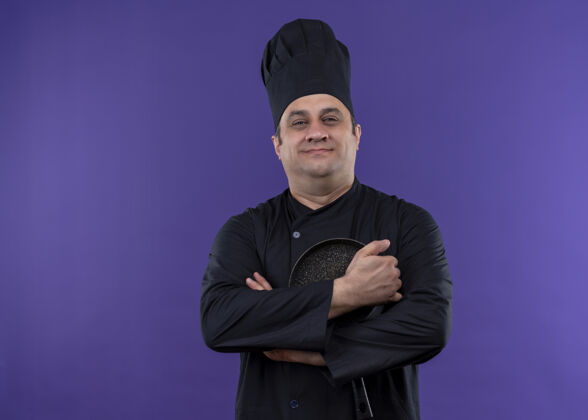 平底锅男厨师身穿黑色制服 头戴厨师帽 手持煎锅 站在紫色背景下 自信地微笑着看着镜头紫色自信制服