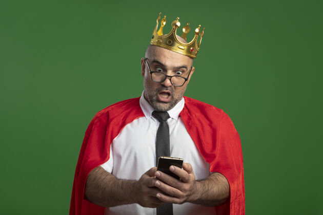 绿色超级英雄商人穿着红斗篷戴着眼镜戴着皇冠用智能手机看着困惑和惊讶斗篷看使用