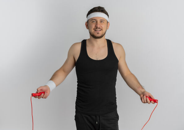 跳跃戴着头带跳绳的年轻健身男子站在白色的背景上 脸上露出幸福的表情健身使用绳索