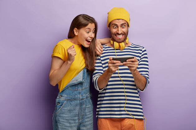 手机兴奋的幸福夫妻用手机玩网络游戏 看智能手机设备 被现代科技看透 穿着时髦的衣服网瘾人耳机牛仔