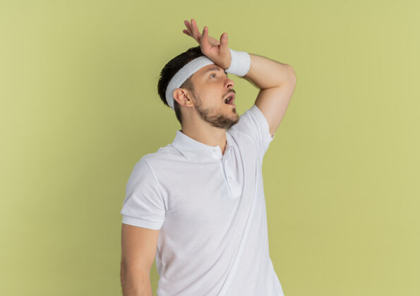 健身身穿白衬衫 头戴头巾的年轻健身男子站在橄榄色背景下 困惑地看着一边 手放在头上手白色旁边