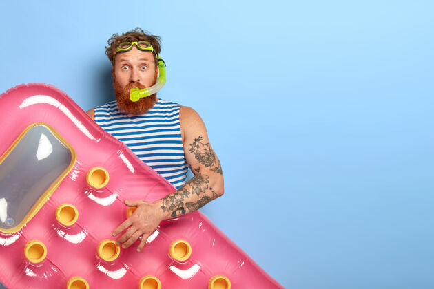 举行卷曲的姜黄色头发的男人戴着泳镜 潜水面罩和充气的粉色床垫 准备在海水中潜水 穿着蓝白相间的条纹背心娱乐护目镜红发