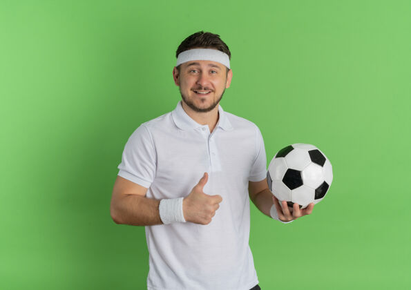 微笑身穿白衬衫 头箍 手持足球 微笑着 竖起大拇指的年轻健身男子站在绿色背景上男士欢呼拇指