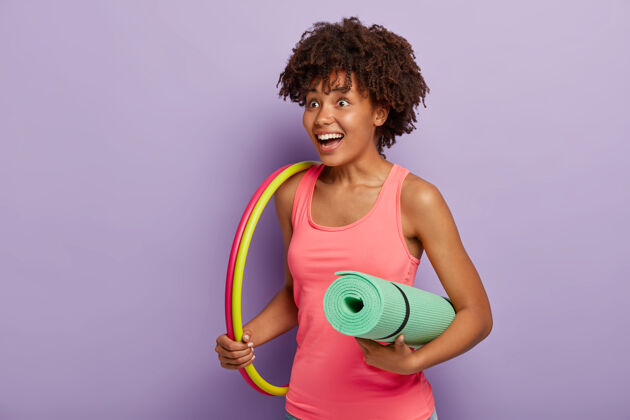 休闲积极的女人留着非洲式发型 拿着卷起的健身垫 带着篮球锻炼 想要保持良好的体形 找个快乐的地方过健康的生活 拿着运动器材粉色紫色女性