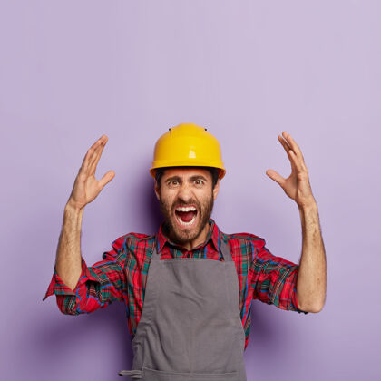 紫色恼怒的情绪化工人戴着黄色的建筑防护头盔 格子衬衫和围裙 有很多工作要做 带着压力和恐慌尖叫 情绪化地举起手臂机械围裙修理