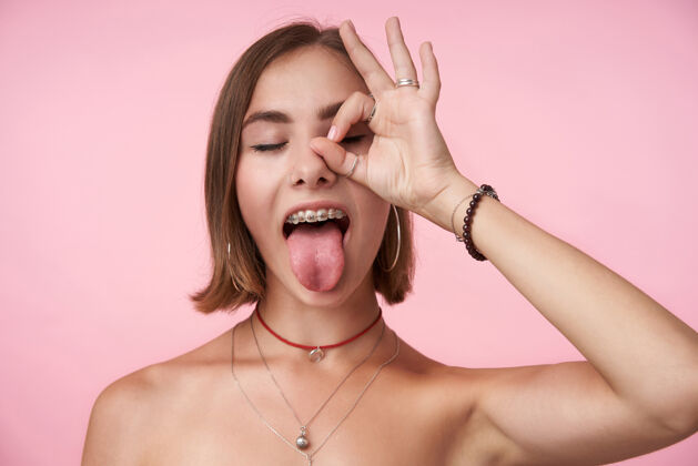 肖像摄影棚拍摄的年轻黑发女性鲍勃剪头发提高ok的姿态 她的脸和伸出舌头 而在粉红色的墙壁上玩的乐趣自然女人穿着