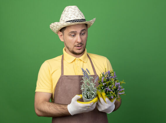 制服年轻的园丁穿着连体衣 戴着帽子 戴着工作手套 手里拿着盆栽植物 站在绿色的墙上看着它们困惑连身衣年轻绿色