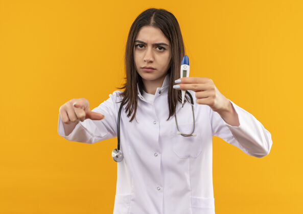 人身穿白大褂的年轻女医生 脖子上戴着听诊器 拿着数字体温计 食指指向前面 严肃的脸站在橙色的墙上外套手势指着
