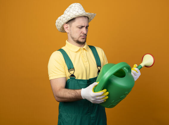 人年轻的园丁穿着连体衣 戴着帽子 手里拿着浇水罐 站在橘色的墙上好奇地看着它花园工作拿着