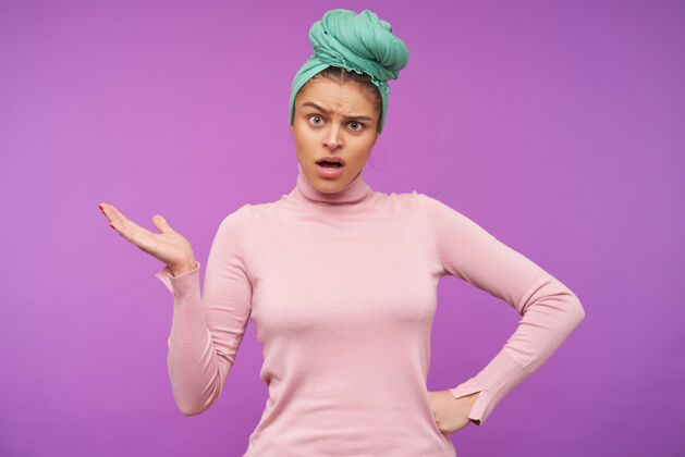发型困惑的年轻优雅的棕色头发的女人困惑地举起她的手掌 同时困惑地看着前面 站在紫色的墙上 戴着头巾和粉色的毛衣自然年轻结